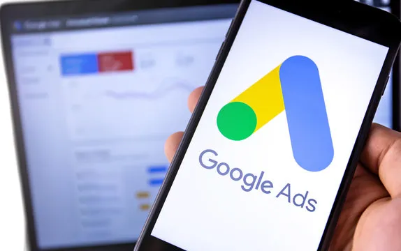 Hand som håller en telefon med Google Ads med Google Ads på desktop i bakgrunden.